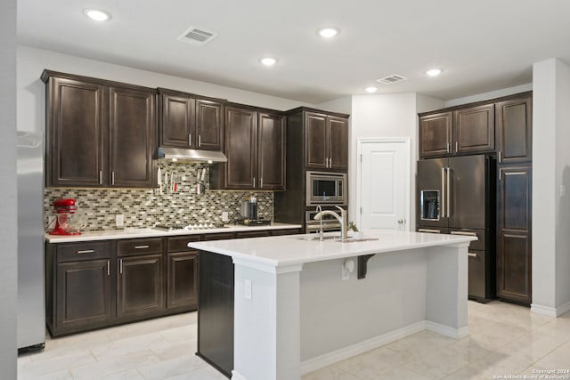 kitchen with backsplash, dark brown cabinets, stainless steel appliances, sink, and a center island with sink