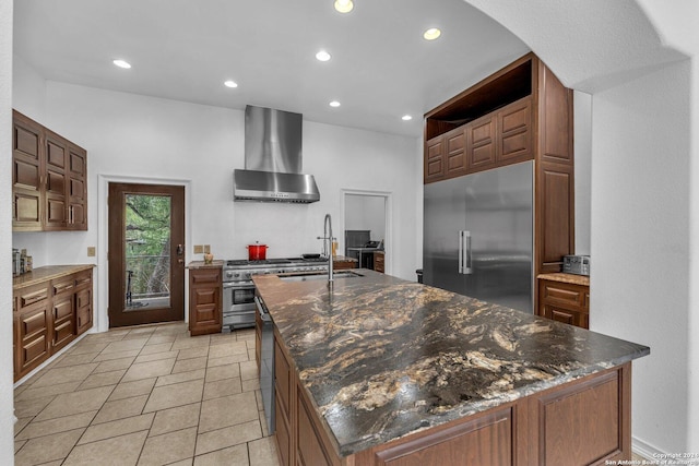 kitchen with sink, wall chimney range hood, dark stone countertops, high quality appliances, and a center island with sink