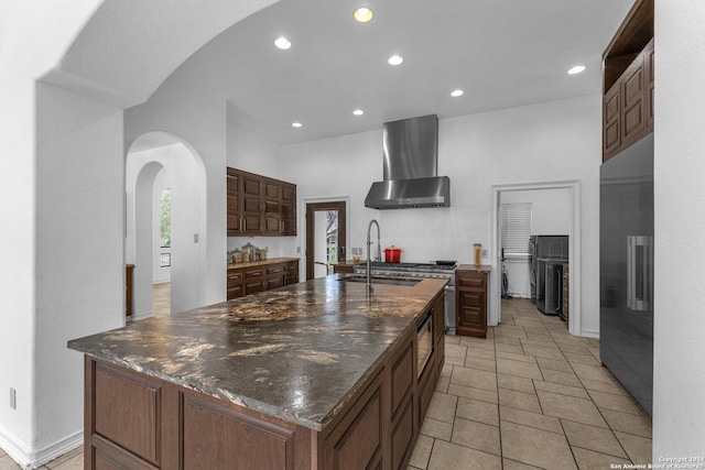 kitchen featuring wall chimney exhaust hood, dark brown cabinets, sink, a center island with sink, and stainless steel refrigerator