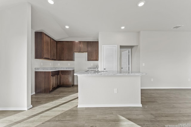 kitchen with lofted ceiling, light stone countertops, light wood-type flooring, an island with sink, and dark brown cabinetry