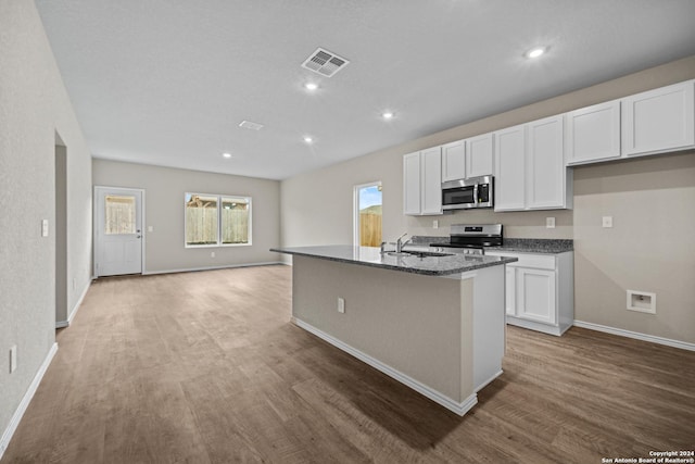 kitchen featuring stainless steel appliances, hardwood / wood-style floors, dark stone counters, a kitchen island with sink, and white cabinets