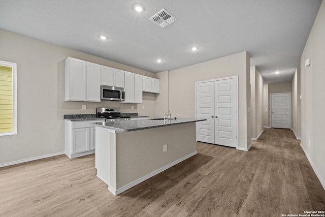 kitchen featuring stainless steel appliances, dark stone countertops, a center island with sink, white cabinets, and light wood-type flooring