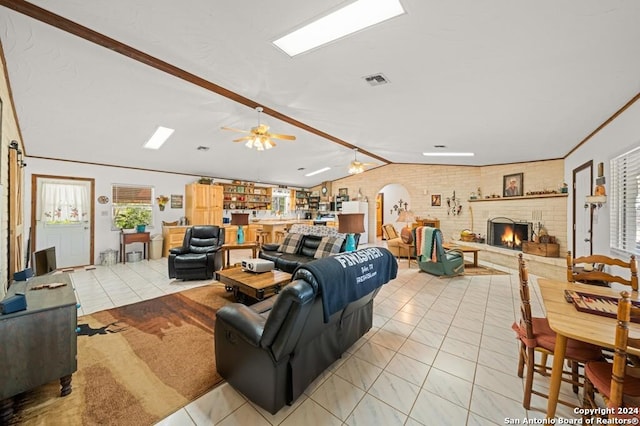 tiled living room with ceiling fan, a brick fireplace, brick wall, vaulted ceiling, and ornamental molding