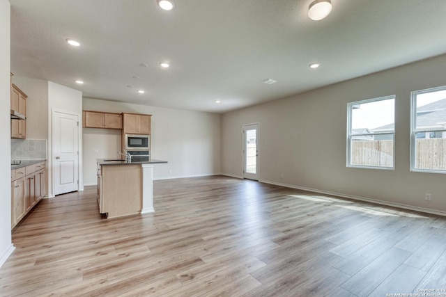 kitchen featuring decorative backsplash, a kitchen island with sink, built in microwave, light brown cabinets, and light hardwood / wood-style floors