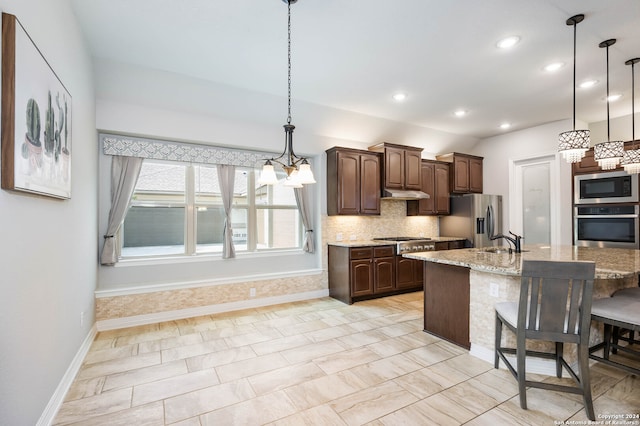 kitchen featuring stainless steel appliances, backsplash, a center island with sink, hanging light fixtures, and light stone countertops