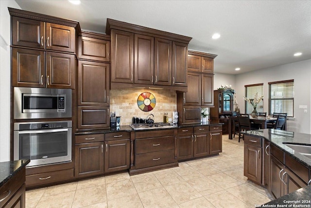 kitchen featuring dark brown cabinetry, backsplash, dark stone countertops, light tile patterned floors, and appliances with stainless steel finishes