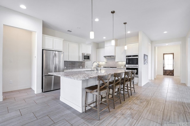 kitchen with a kitchen island with sink, white cabinets, hanging light fixtures, light stone counters, and stainless steel appliances