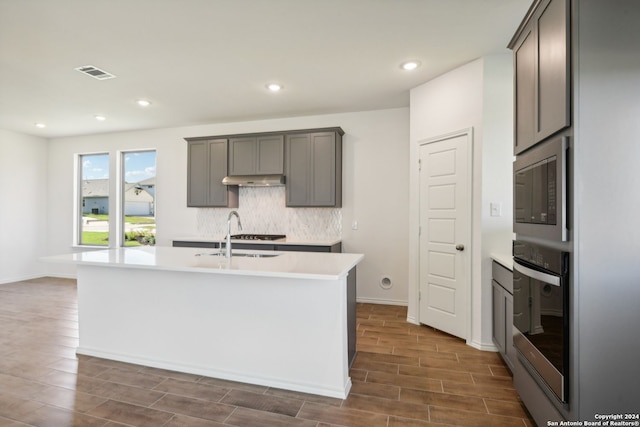 kitchen with sink, gray cabinetry, stainless steel appliances, a center island with sink, and decorative backsplash