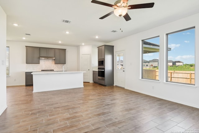 kitchen featuring appliances with stainless steel finishes, backsplash, a kitchen island with sink, ceiling fan, and light hardwood / wood-style floors