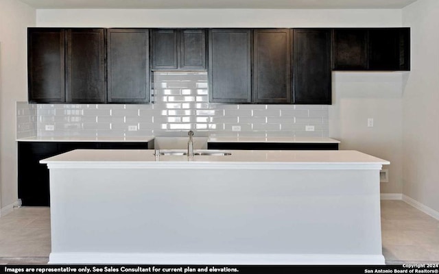 kitchen with a kitchen island with sink, dark brown cabinets, backsplash, sink, and light tile flooring