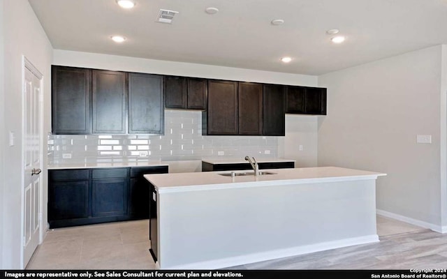 kitchen featuring dark brown cabinetry, light tile flooring, backsplash, an island with sink, and sink