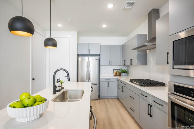 kitchen with wall chimney range hood, hanging light fixtures, sink, appliances with stainless steel finishes, and light hardwood / wood-style floors