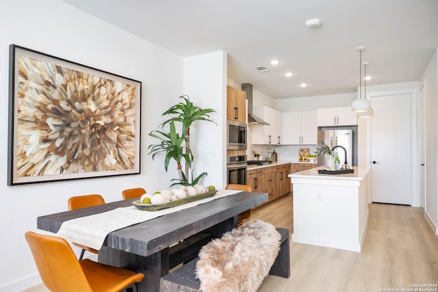 kitchen with wall chimney exhaust hood, light hardwood / wood-style floors, pendant lighting, a kitchen island with sink, and appliances with stainless steel finishes