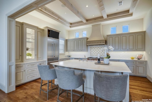 kitchen with custom exhaust hood, stainless steel built in refrigerator, a kitchen island with sink, and hardwood / wood-style floors