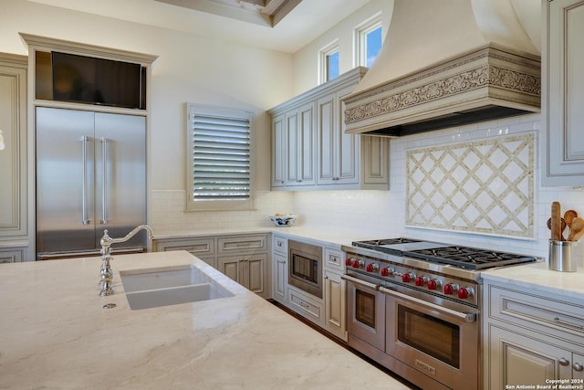 kitchen with sink, light stone counters, built in appliances, custom range hood, and decorative backsplash