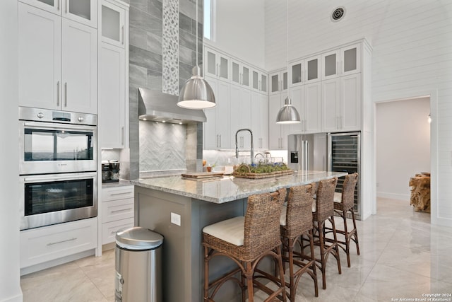 kitchen with light stone countertops, a high ceiling, a center island with sink, white cabinets, and appliances with stainless steel finishes