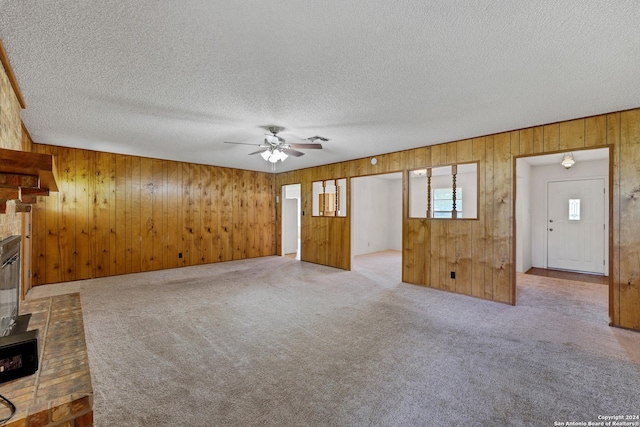 unfurnished living room featuring a brick fireplace, a textured ceiling, wooden walls, carpet floors, and ceiling fan