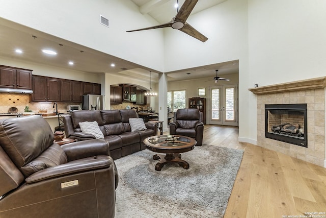 living room with ceiling fan, beam ceiling, high vaulted ceiling, a tiled fireplace, and light wood-type flooring