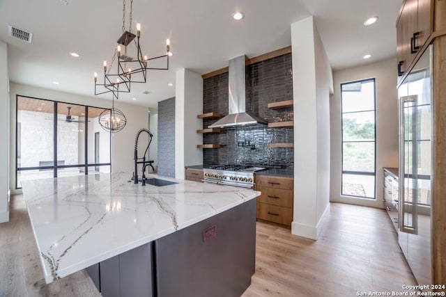 kitchen with wall chimney range hood, sink, light stone counters, decorative light fixtures, and a large island with sink