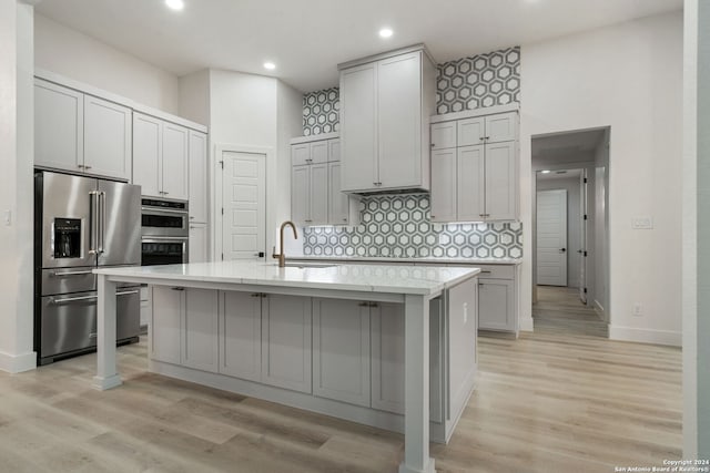 kitchen with decorative backsplash, appliances with stainless steel finishes, sink, light hardwood / wood-style floors, and an island with sink