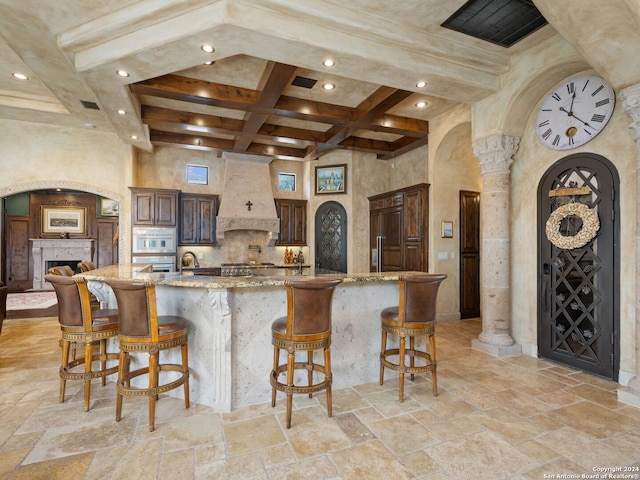 kitchen with light stone counters, custom range hood, a kitchen bar, and coffered ceiling