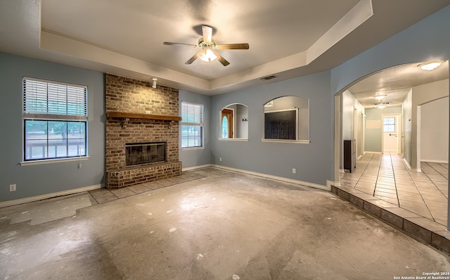 unfurnished living room with a tray ceiling, ceiling fan, a fireplace, and plenty of natural light