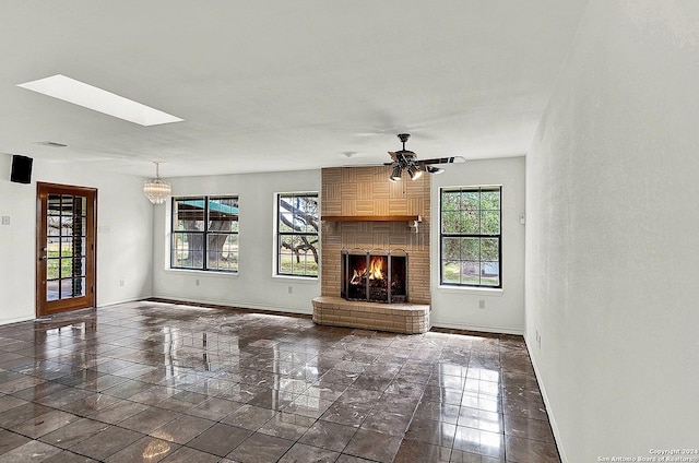 unfurnished living room with ceiling fan with notable chandelier, a skylight, and a brick fireplace