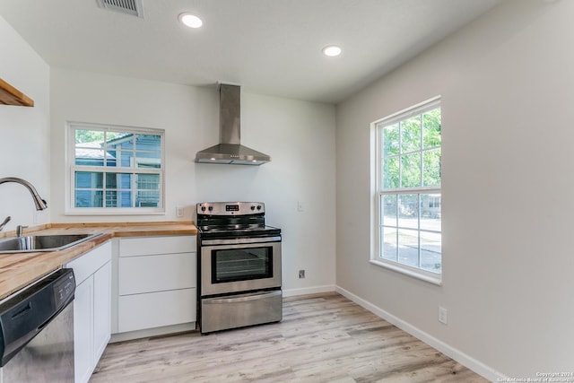 kitchen featuring white cabinets, wall chimney range hood, sink, appliances with stainless steel finishes, and butcher block countertops