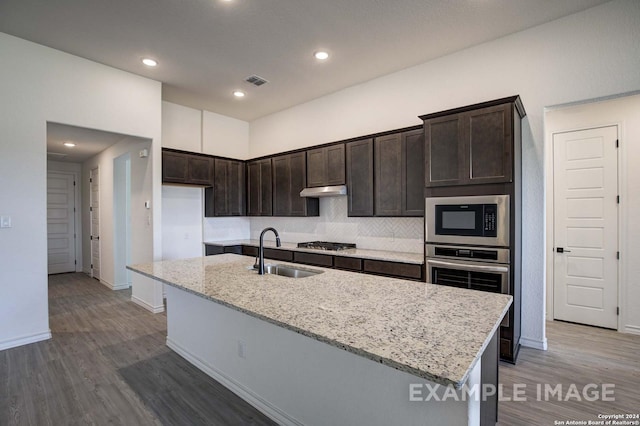 kitchen featuring appliances with stainless steel finishes, a kitchen island with sink, wood-type flooring, light stone counters, and sink