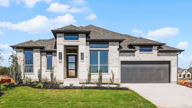 prairie-style home with roof with shingles, driveway, an attached garage, a front yard, and brick siding