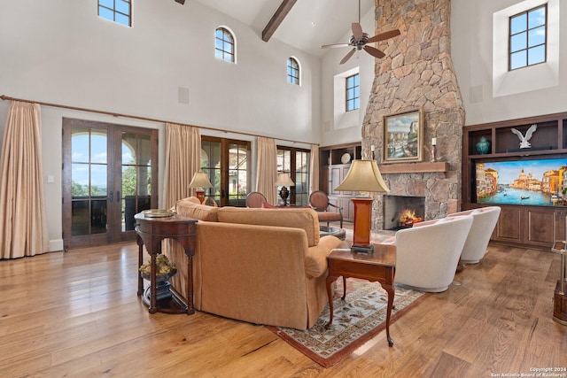 living room with french doors, ceiling fan, beamed ceiling, a high ceiling, and a stone fireplace