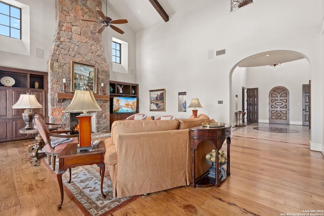 living room with a towering ceiling, ceiling fan, beamed ceiling, a stone fireplace, and plenty of natural light