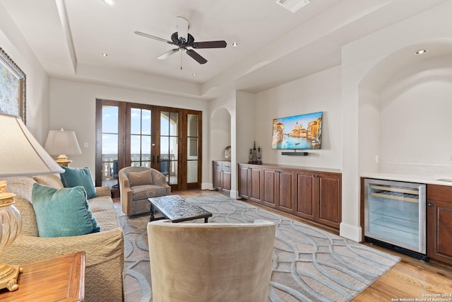 living room featuring french doors, a tray ceiling, beverage cooler, ceiling fan, and light hardwood / wood-style flooring