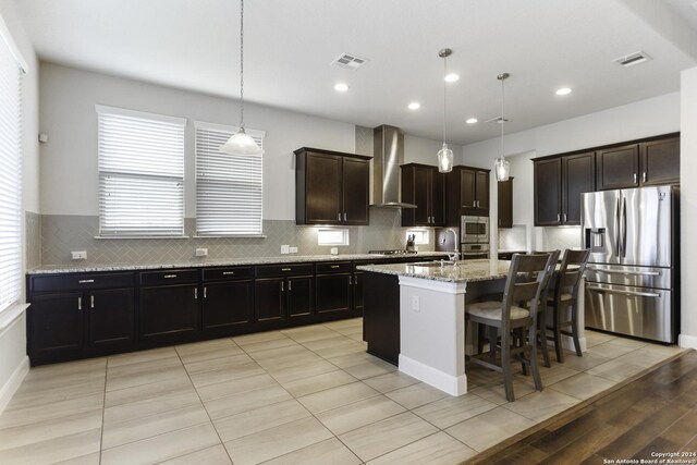 kitchen featuring light tile patterned floors, decorative light fixtures, stainless steel appliances, a center island with sink, and wall chimney exhaust hood