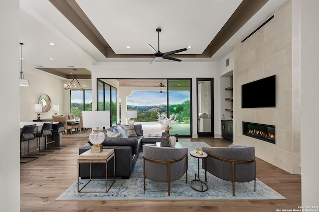 living room with a tile fireplace, ceiling fan with notable chandelier, hardwood / wood-style flooring, built in features, and a tray ceiling