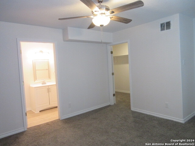 unfurnished bedroom with a closet, ensuite bathroom, ceiling fan, dark carpet, and a spacious closet