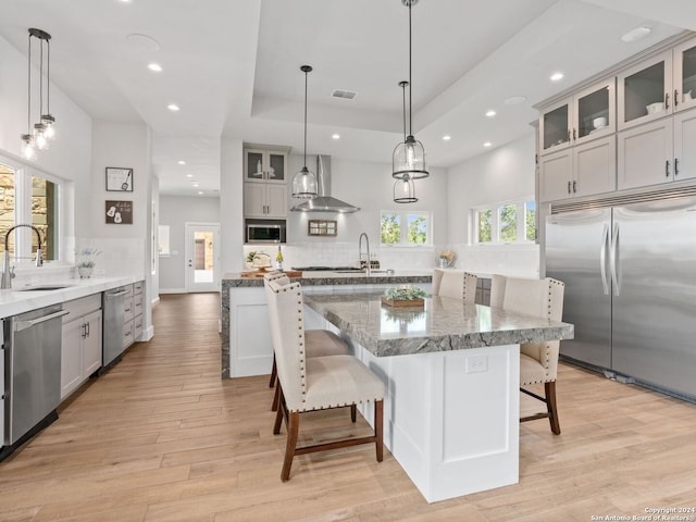 kitchen with wall chimney range hood, built in appliances, light hardwood / wood-style floors, and decorative light fixtures