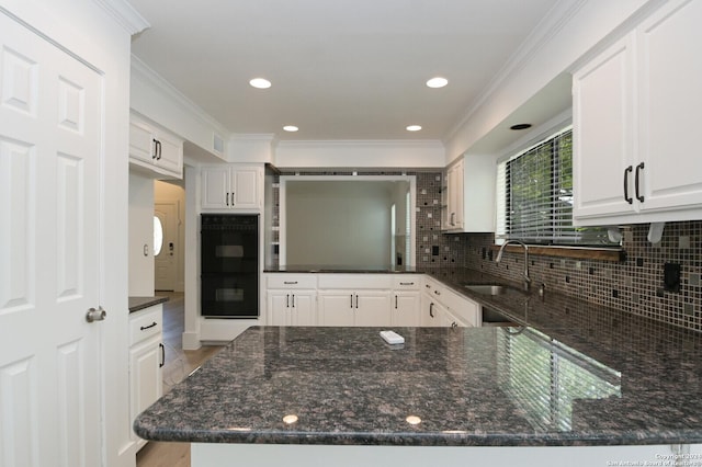 kitchen with kitchen peninsula, decorative backsplash, dark stone countertops, double oven, and white cabinetry