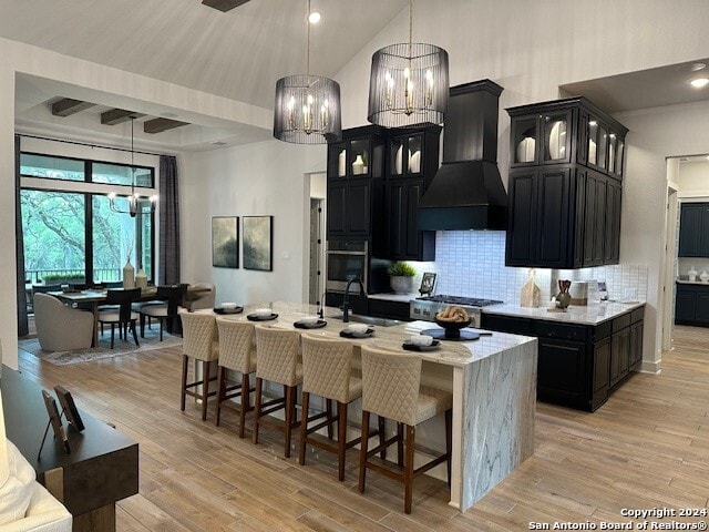 kitchen with decorative light fixtures, backsplash, a center island with sink, sink, and custom range hood