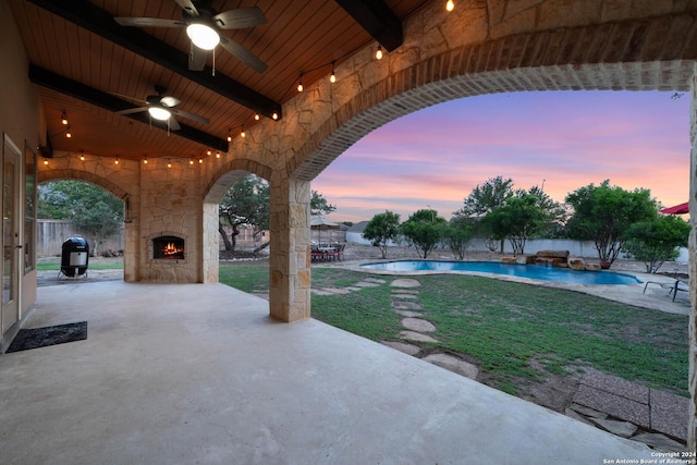 patio terrace at dusk with ceiling fan, a fenced in pool, area for grilling, and an outdoor stone fireplace