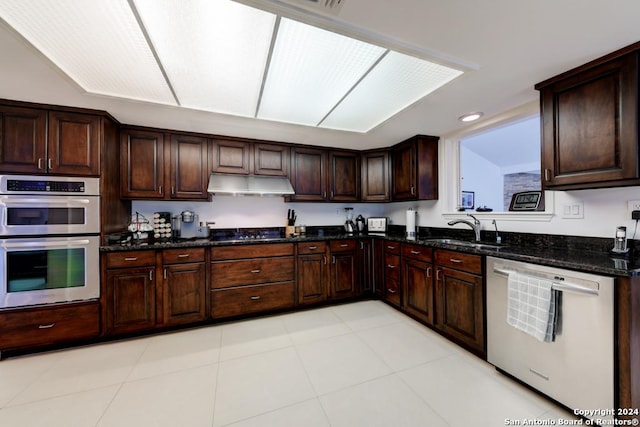 kitchen with appliances with stainless steel finishes, light tile patterned flooring, dark stone countertops, dark brown cabinetry, and sink