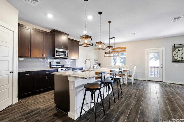 kitchen featuring sink, appliances with stainless steel finishes, hanging light fixtures, light stone countertops, and a center island with sink