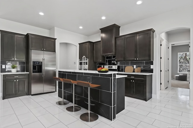 kitchen with dark brown cabinets, stainless steel refrigerator with ice dispenser, a center island with sink, and decorative backsplash