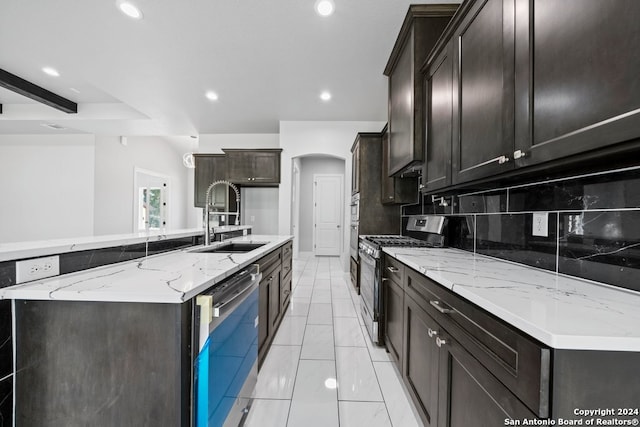 kitchen with a kitchen island with sink, sink, decorative backsplash, stainless steel appliances, and light stone countertops