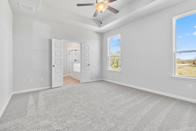 unfurnished bedroom featuring a raised ceiling, ensuite bath, ceiling fan, and light colored carpet