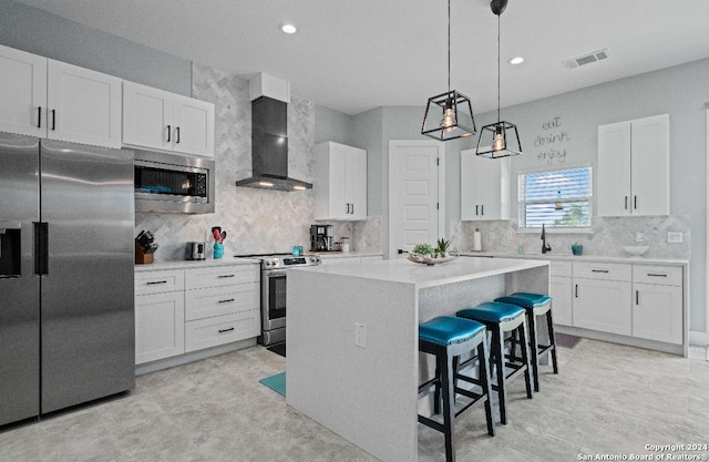 kitchen featuring hanging light fixtures, wall chimney exhaust hood, stainless steel appliances, white cabinets, and a kitchen island