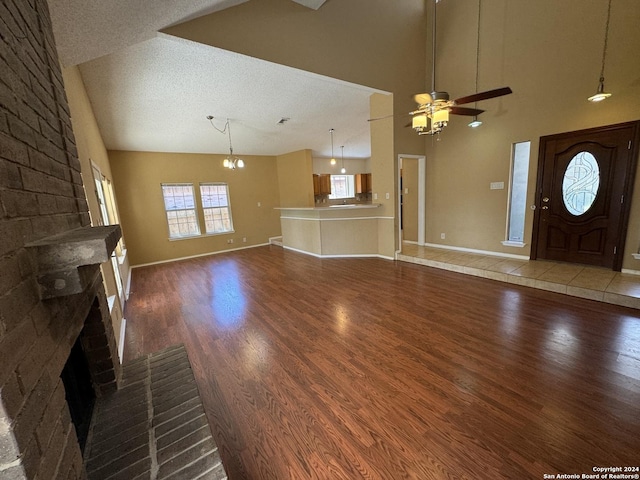 unfurnished living room featuring dark hardwood / wood-style floors, ceiling fan with notable chandelier, high vaulted ceiling, a brick fireplace, and a textured ceiling