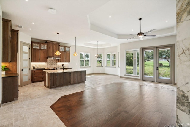 kitchen featuring tasteful backsplash, hanging light fixtures, a raised ceiling, light stone countertops, and a center island with sink