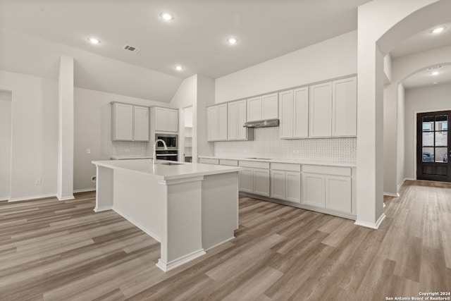 kitchen with backsplash, vaulted ceiling, an island with sink, light hardwood / wood-style floors, and stainless steel microwave