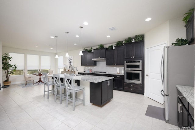 kitchen featuring light stone countertops, a kitchen breakfast bar, stainless steel appliances, decorative light fixtures, and an island with sink